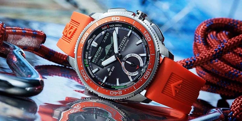 Een sportief Citizen Eco-Drive horloge met oranje band ligt op een reflecterend oppervlak naast nautische touwen en beslag.