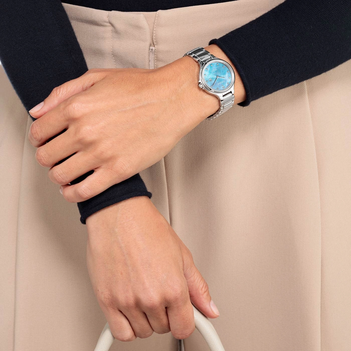 Vrouw met zilveren horloge met blauwe wijzerplaat, houdt een witte tas vast.