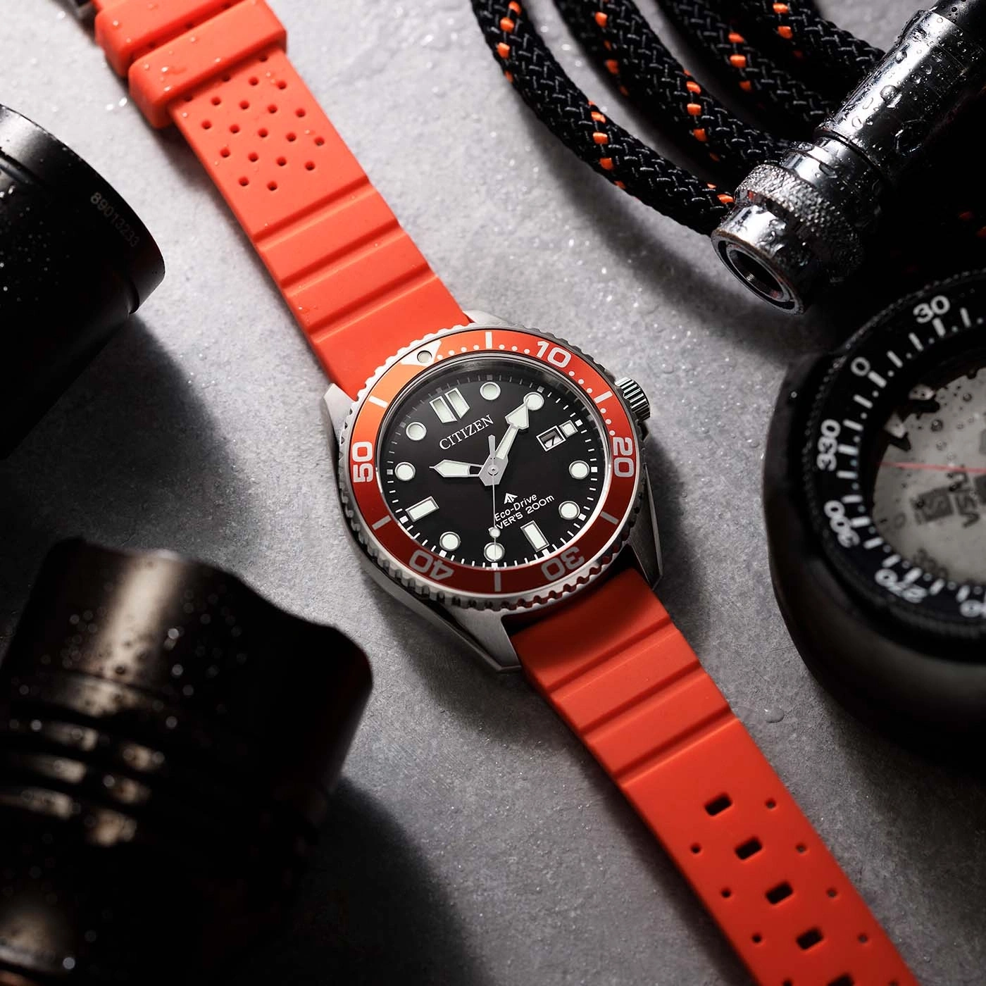 Close-up van een Citizen Eco-Drive duikhorloge met rode band, naast een kompas en zaklamp.