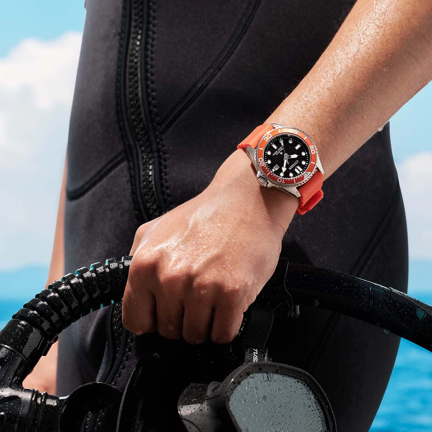 Een persoon in een wetsuit houdt duikuitrusting vast, met een Citizen horloge om.