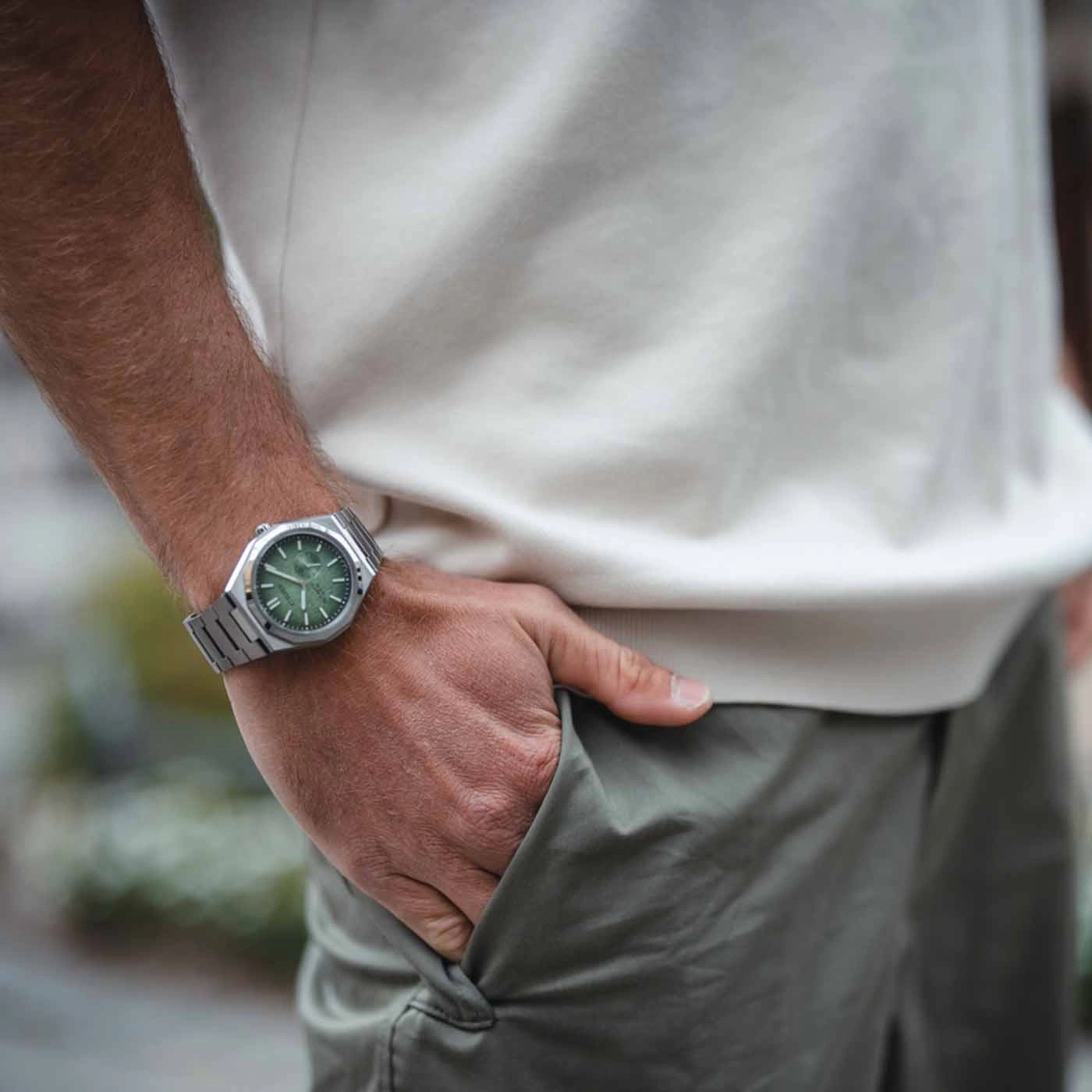 Man draagt zilveren horloge met groene wijzerplaat, hand in zak.