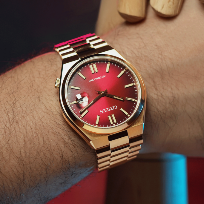 Een automatisch horloge van Citizen met een rode wijzerplaat en goudkleurige afwerking wordt getoond op een harige pols. Het horloge staat centraal.
