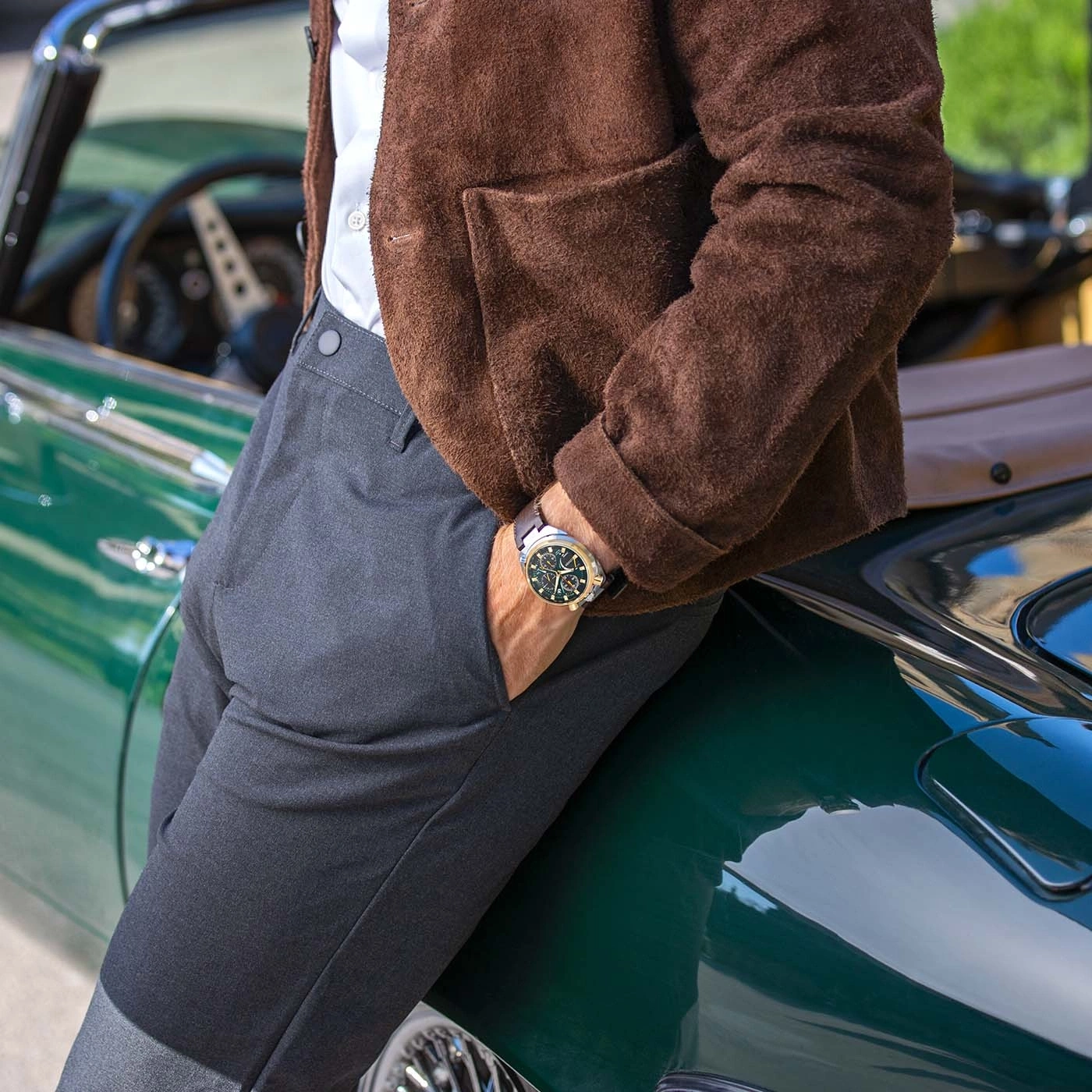 Man leunt tegen een vintage groene auto, draagt een bruine jas en een horloge.
