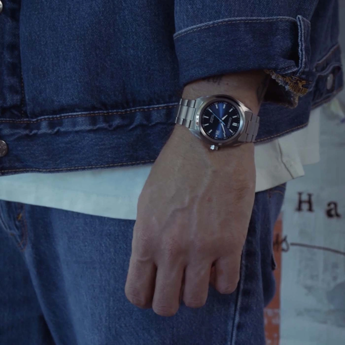 Man draagt een blauw spijkerjack, wit T-shirt en spijkerbroek met horloge