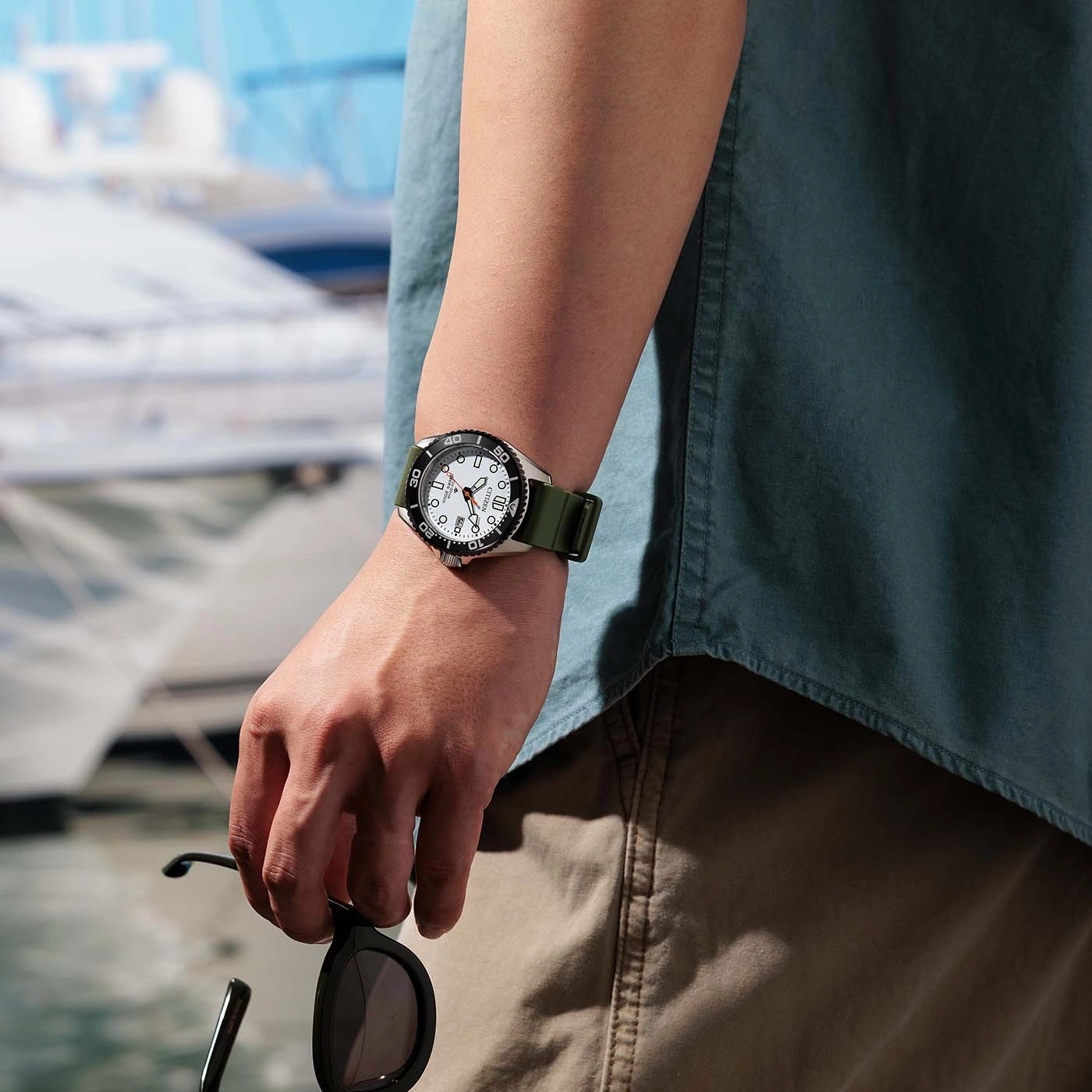 Man met horloge en zonnebril aan het water met boten op de achtergrond.