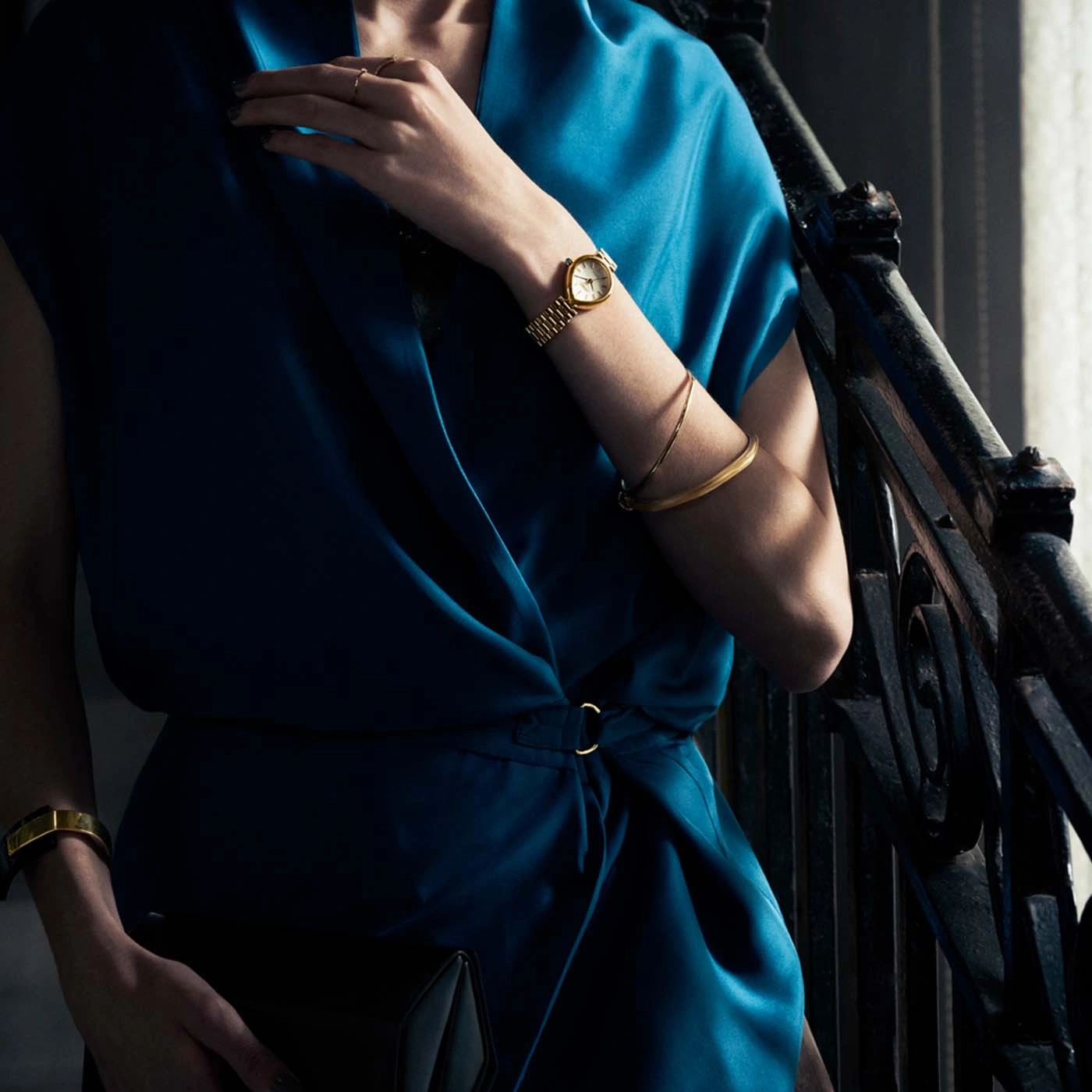 Vrouw in blauwe jurk met gouden sieraden en clutch.