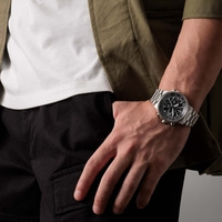 Man met horloge, zwarte broek, wit shirt en groene jas.
