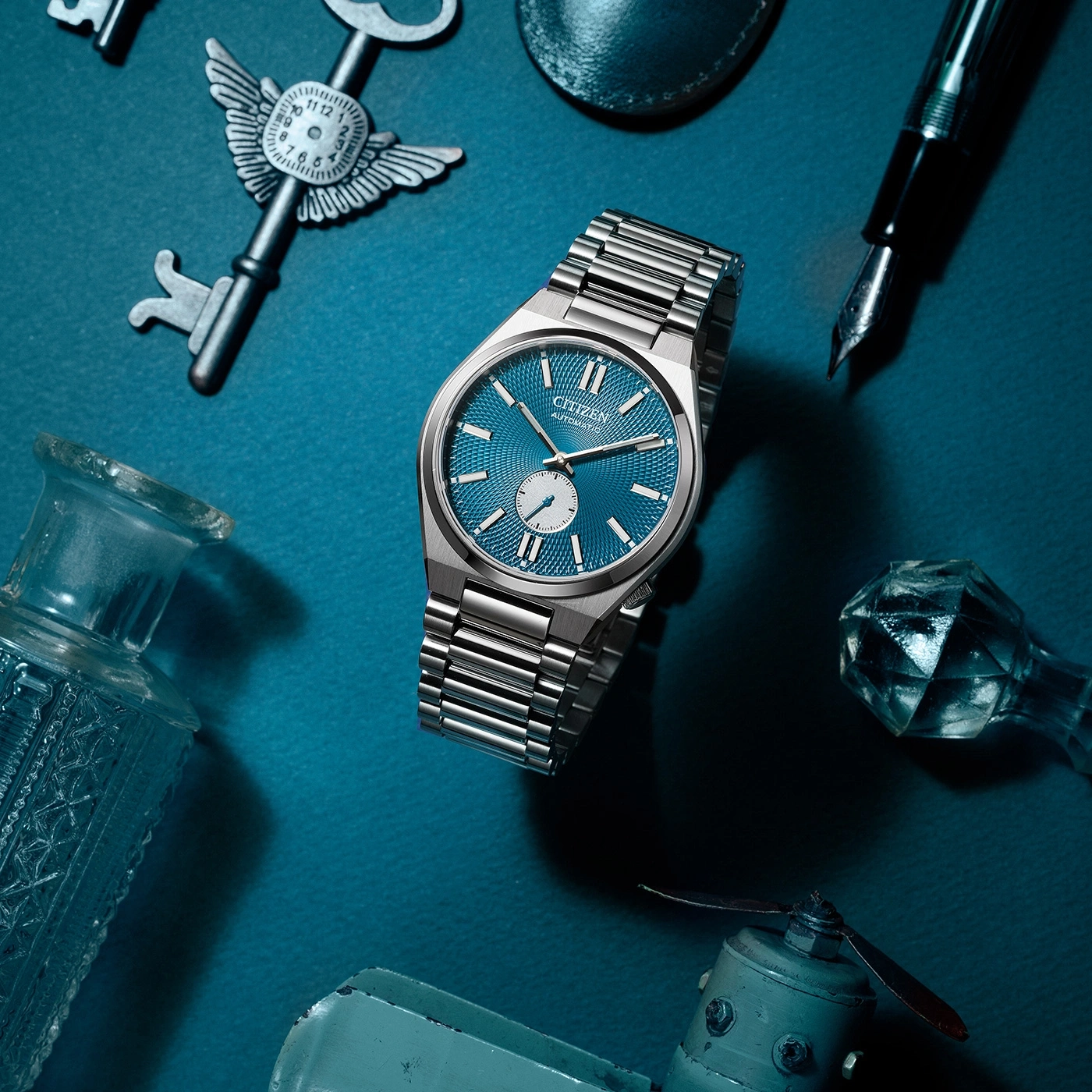 Citizen horloge met blauwe wijzerplaat, sleutels, pen en andere objecten op blauwe ondergrond.