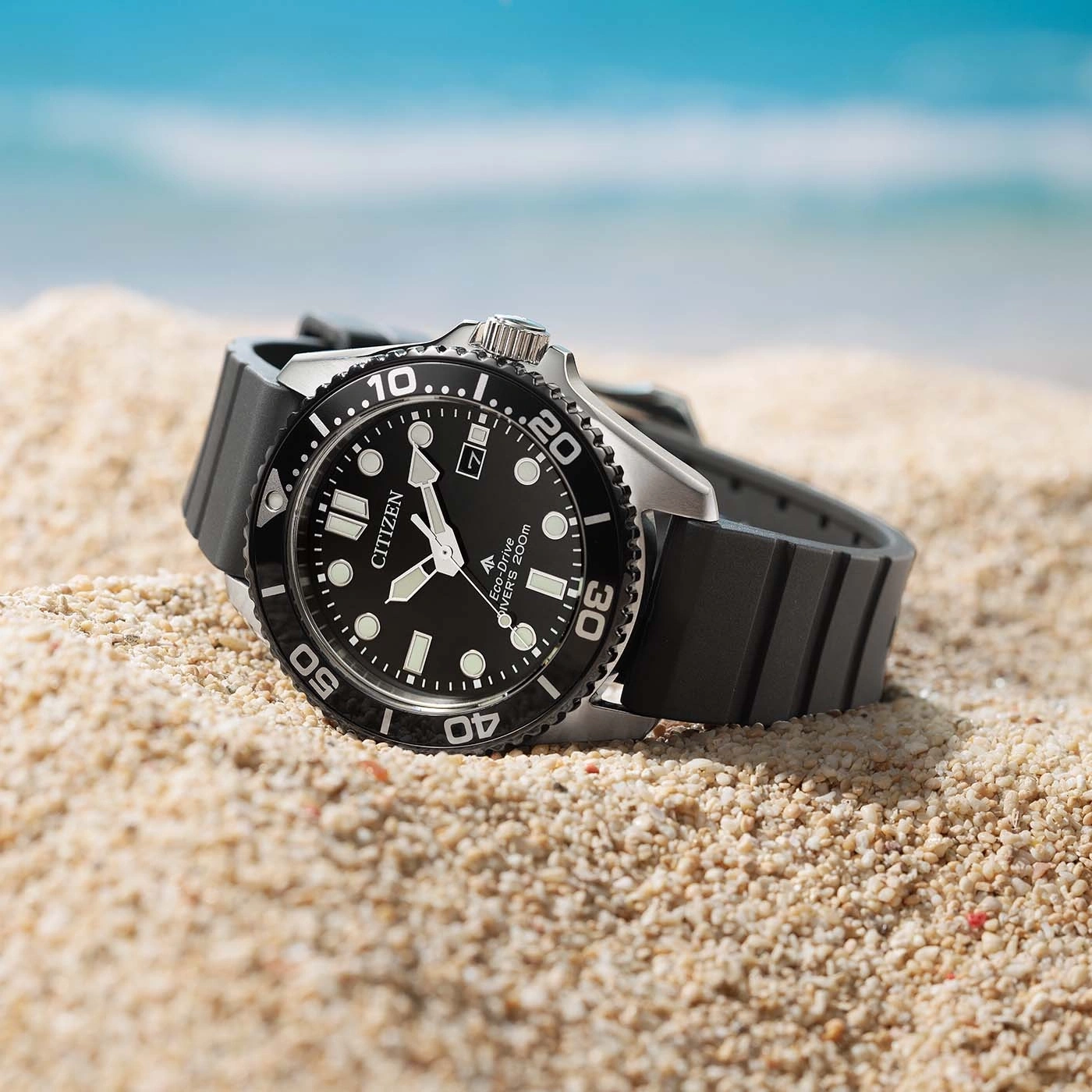 Een zwart Citizen Eco-Drive duikhorloge ligt op een zandstrand met oceaan op de achtergrond.