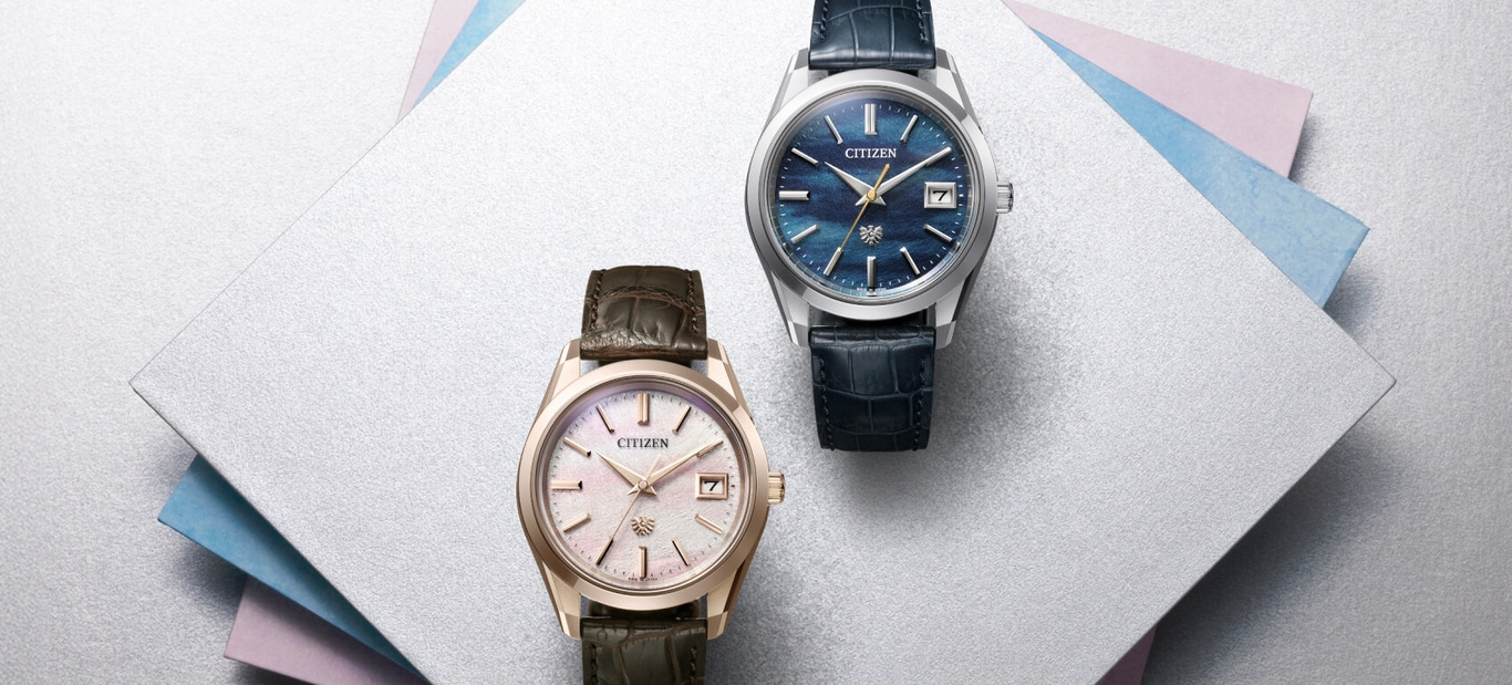 Twee Citizen horloges op een zilveren oppervlak met kleurrijke papieren accenten.