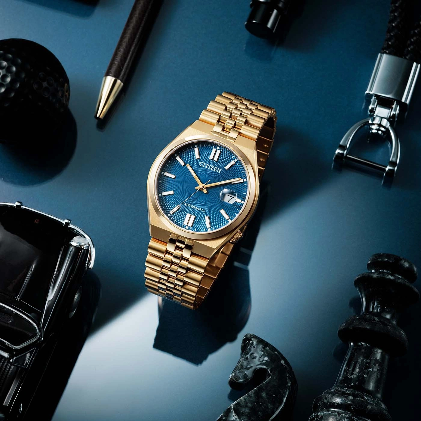 Gouden horloge met blauwe wijzerplaat, omringd door accessoires op blauwe ondergrond.