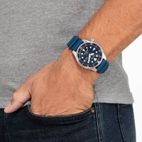 Man met een Citizen horloge met blauwe band, hand in de zak, jeans en grijs shirt.