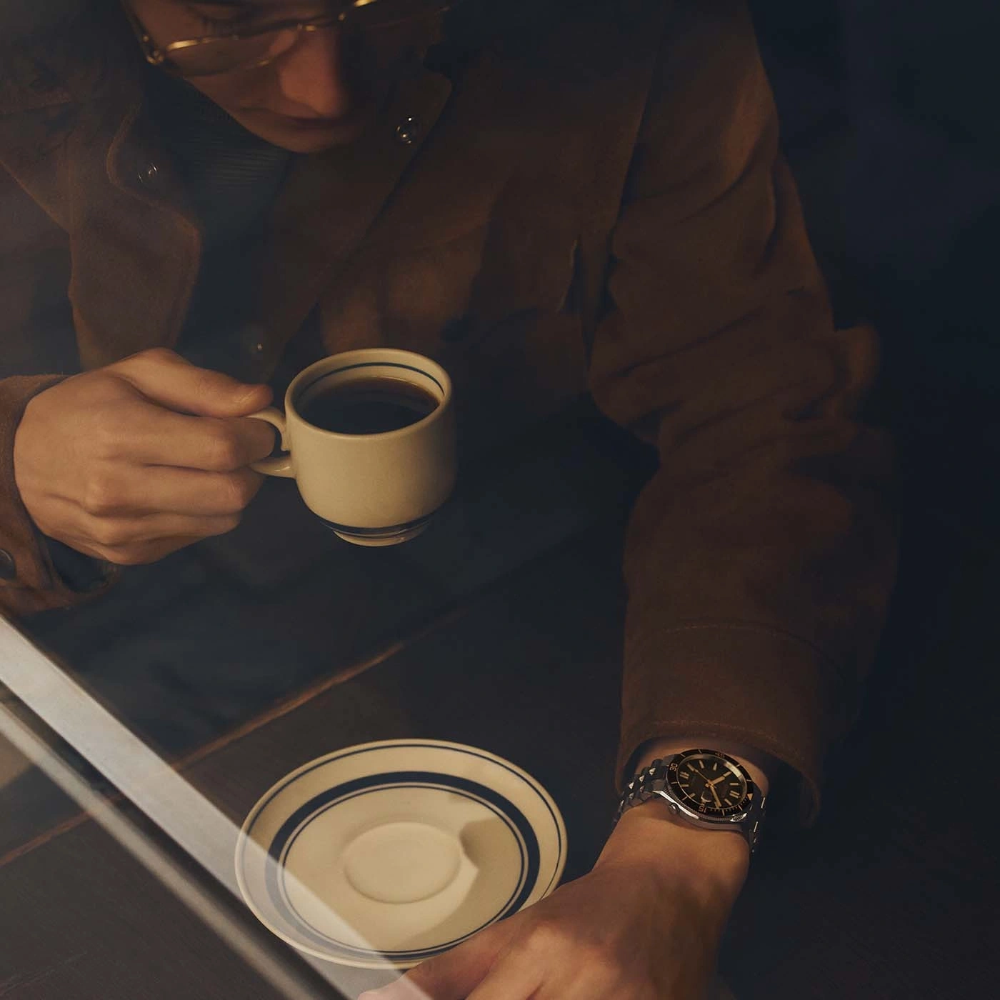 Man in bruine jas drinkt koffie, draagt een horloge.