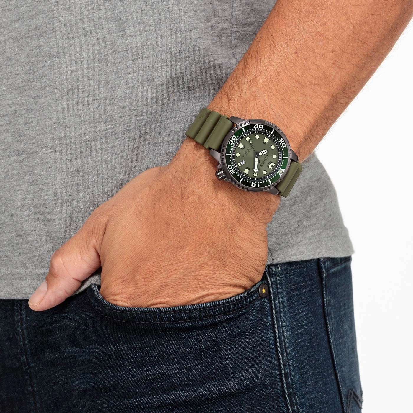 Man met groen horloge, hand in zak, grijs shirt, blauwe jeans.
