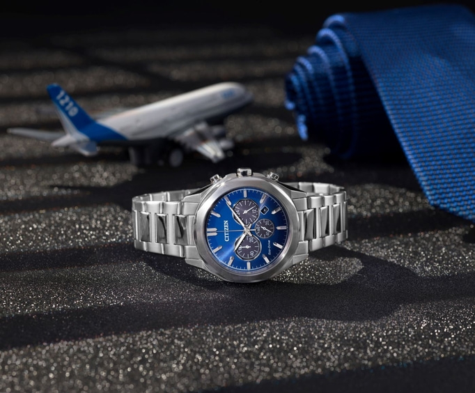 Een Citizen-horloge met een blauwe wijzerplaat is prominent aanwezig, samen met een vliegtuigmodel en een blauwe stropdas, allemaal gepresenteerd op een sprankelend, donker oppervlak.
